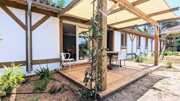 A modern outdoor patio with wooden beams and a pergola, surrounded by a garden with plants and a small table with chairs. - Jasmin - Nature & sea with terrace and play area (Lesperon)