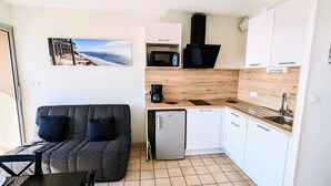 A compact kitchenette with a microwave, coffee maker, and a small refrigerator. There's a wooden countertop and a sink. A patterned sofa is against the wall with a beach photo above it. - Appartement T2 Front de mer (Mimizan)