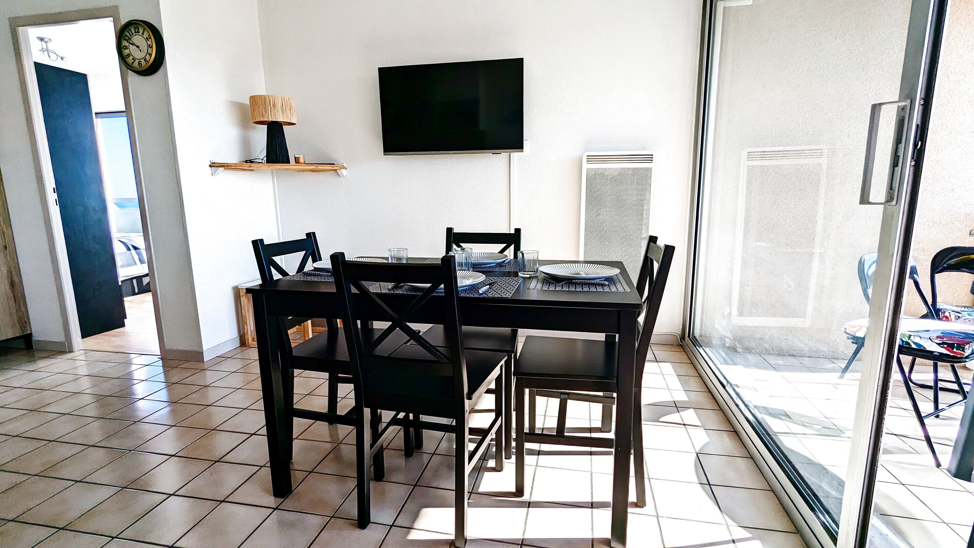Un coin repas avec une table dressée pour quatre personnes, des chaises, un téléviseur fixé au mur et une horloge.