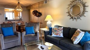 Una sala de estar con una mesa de centro de vidrio, dos sillones, un sofá a rayas, un espejo decorativo y una lámpara.