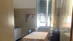 Un petit coin repas avec une table et des chaises, une fenêtre avec des stores et, au fond, un espace cuisine.