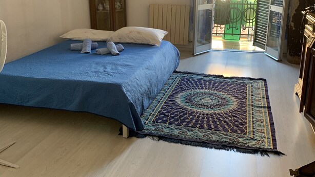 Une chambre à coucher avec un lit, un radiateur, une plante en pot et une vue sur l’extérieur, visible à travers une porte ouverte.