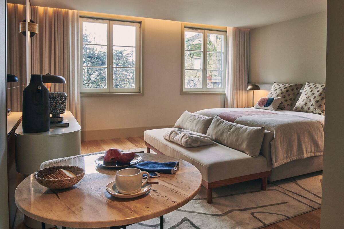 Habitación de hotel con una cama, una mesa redonda con un cuenco y una taza, y dos ventanas con cortinas.