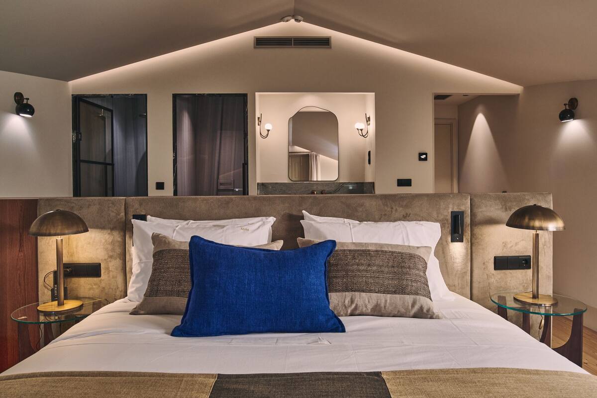 Una habitación de hotel moderna con una cama grande, dos lámparas de noche, un espejo sobre la cama y una mesita con una lámpara.