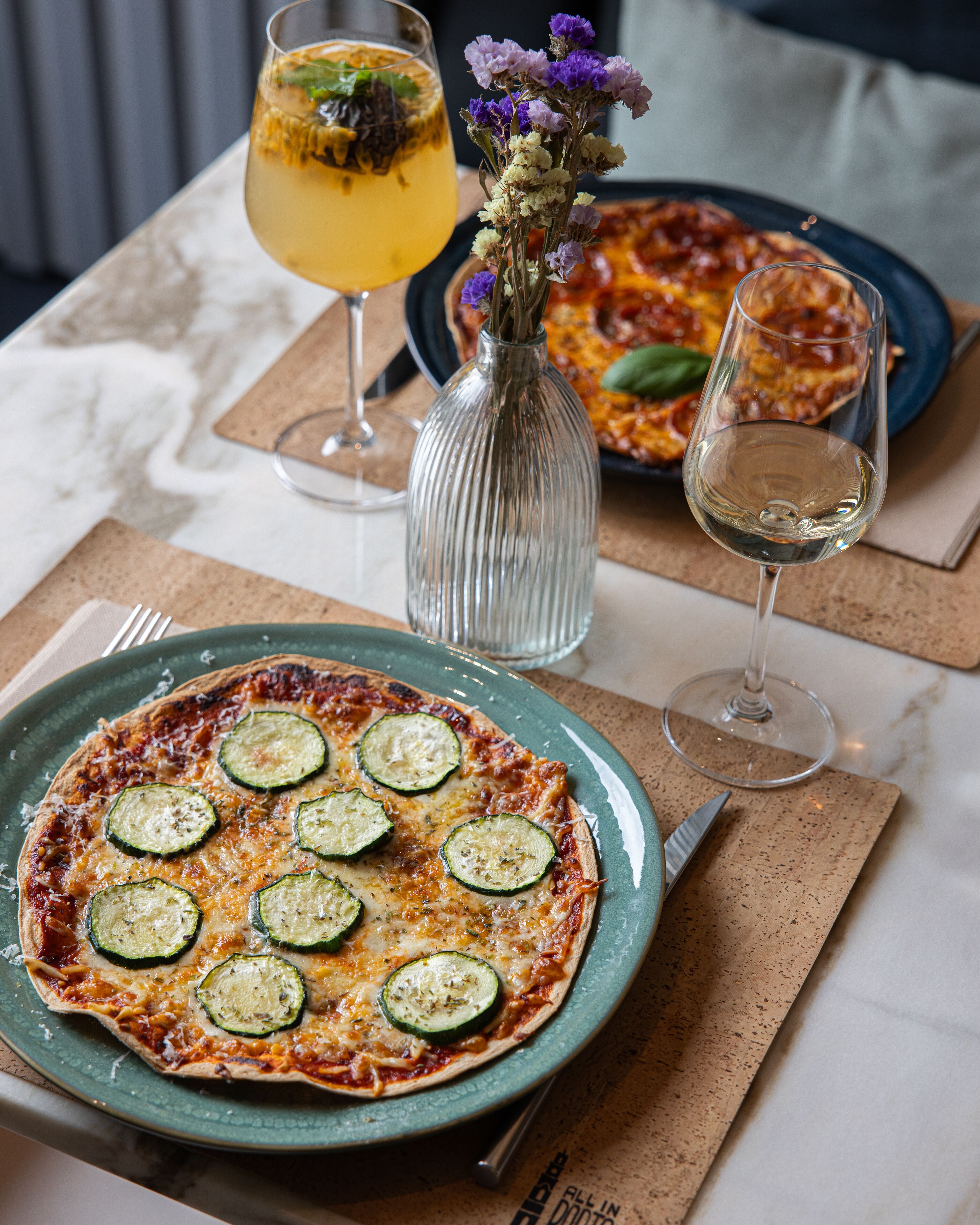 A table with a pizza, a glass of white wine, and a glass of yellow drink.