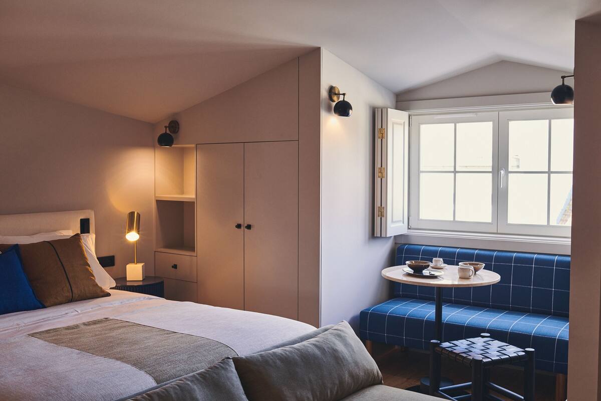 Habitación de hotel con cama, mesita de noche con lámpara, armario, ventana con vistas y una pequeña mesa con un banco.