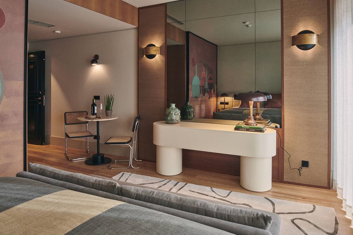 Un dormitorio moderno con una cama grande, una mesita de noche, un espejo y un pequeño rincón de descanso con una silla y una mesa.