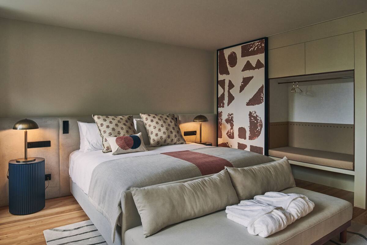 Una habitación de hotel moderna con una cama grande, mesitas de noche, un banco y una pared con arte abstracto.