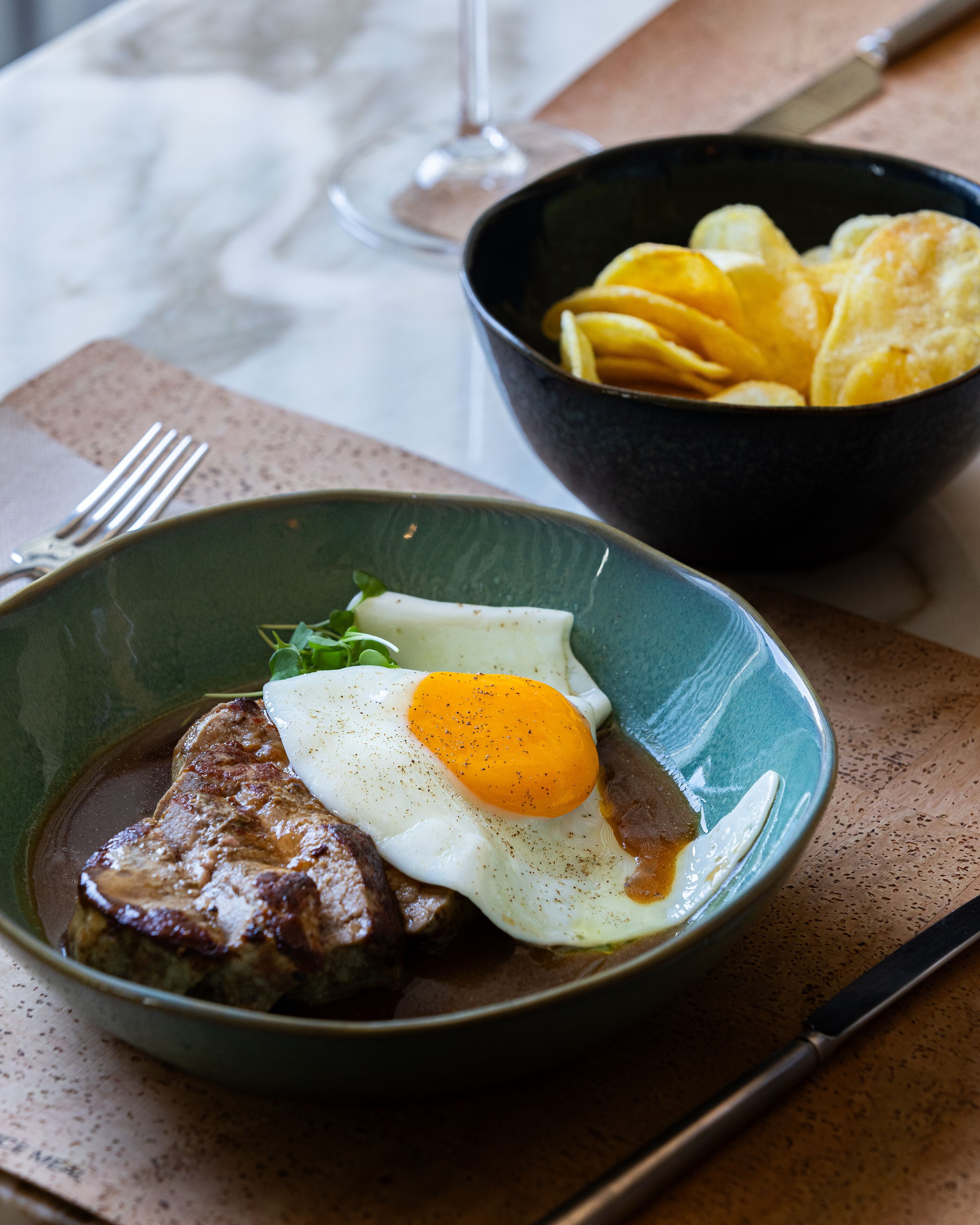 A plate with steak and a fried egg, a bowl of chips, and a fork on a table.
