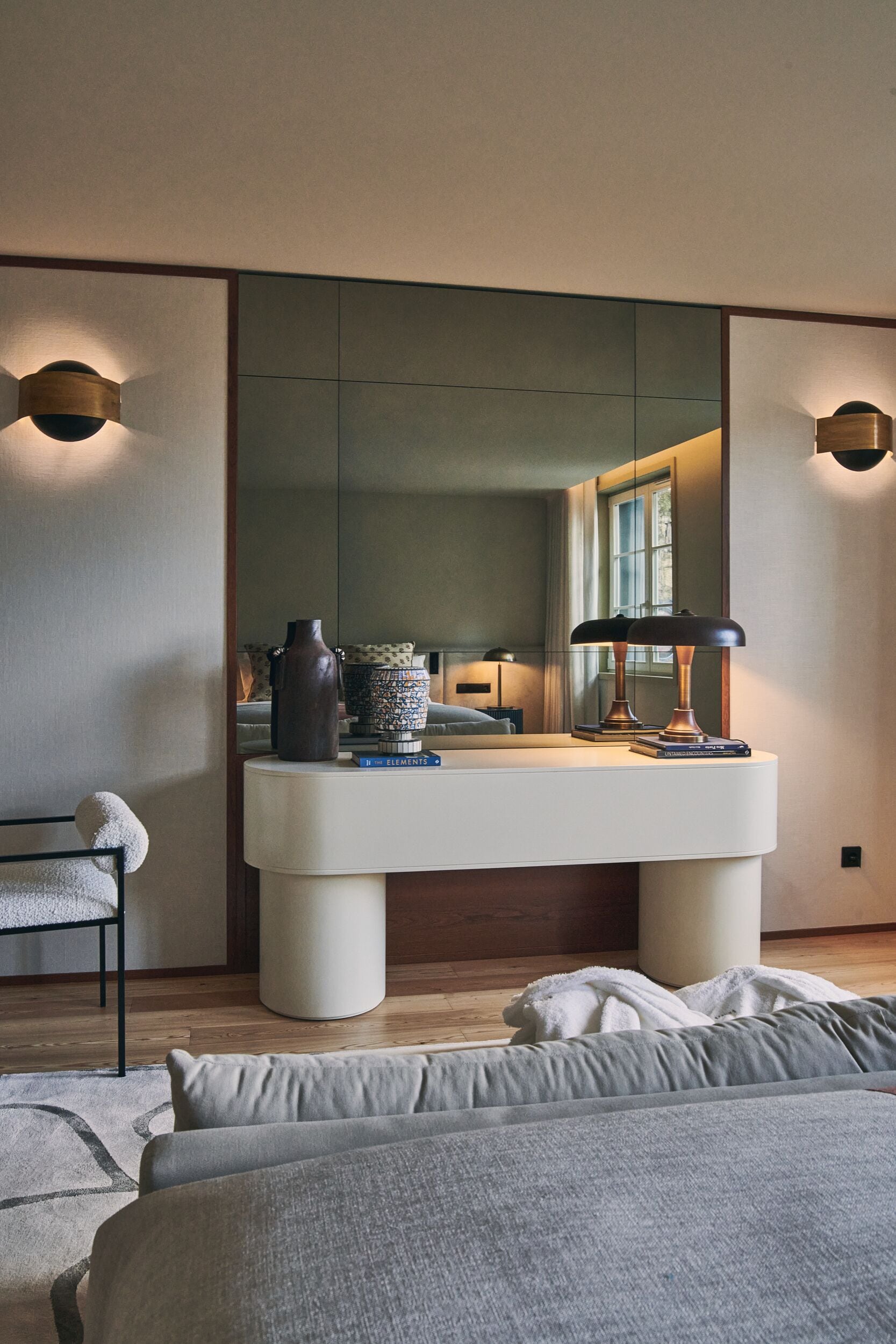 A modern bedroom with a large bed, a bedside table with a lamp, a mirror, and a window with curtains.