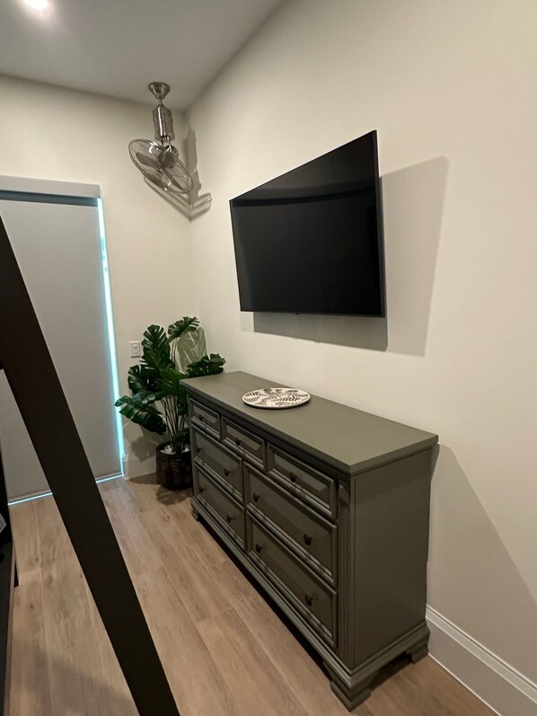 A modern living room with a flat-screen TV mounted on the wall, a wooden floor, a dark-colored cabinet with drawers, a potted plant, and a ceiling fan.