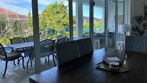 Un'area patio moderna con tavolo, sedie e panchina. Sono presenti ampie porte in vetro che danno su una piscina e una vista di una zona residenziale con alberi e case.