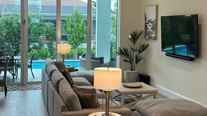 Un soggiorno spazioso con un divano ampio, un tavolino e un televisore montato a parete. È presente un ventilatore a soffitto e una finestra con vista su una piscina e vegetazione.