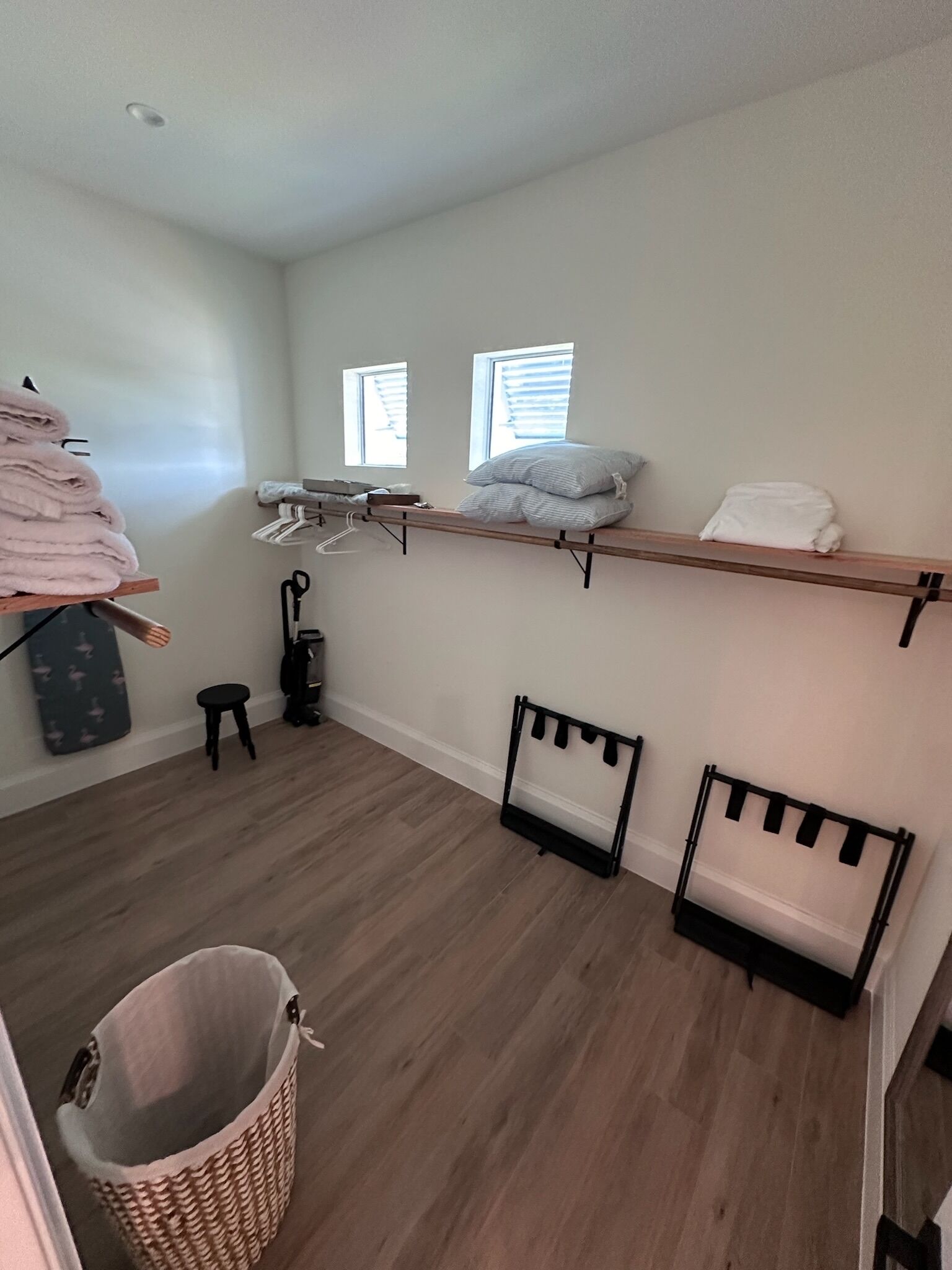 Una habitación con piso de madera, un cesto de ropa, un taburete pequeño y un estante con toallas dobladas y almohadas.