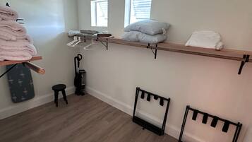 Una habitación con piso de madera, un cesto de ropa, un taburete pequeño y un estante con toallas dobladas y almohadas.