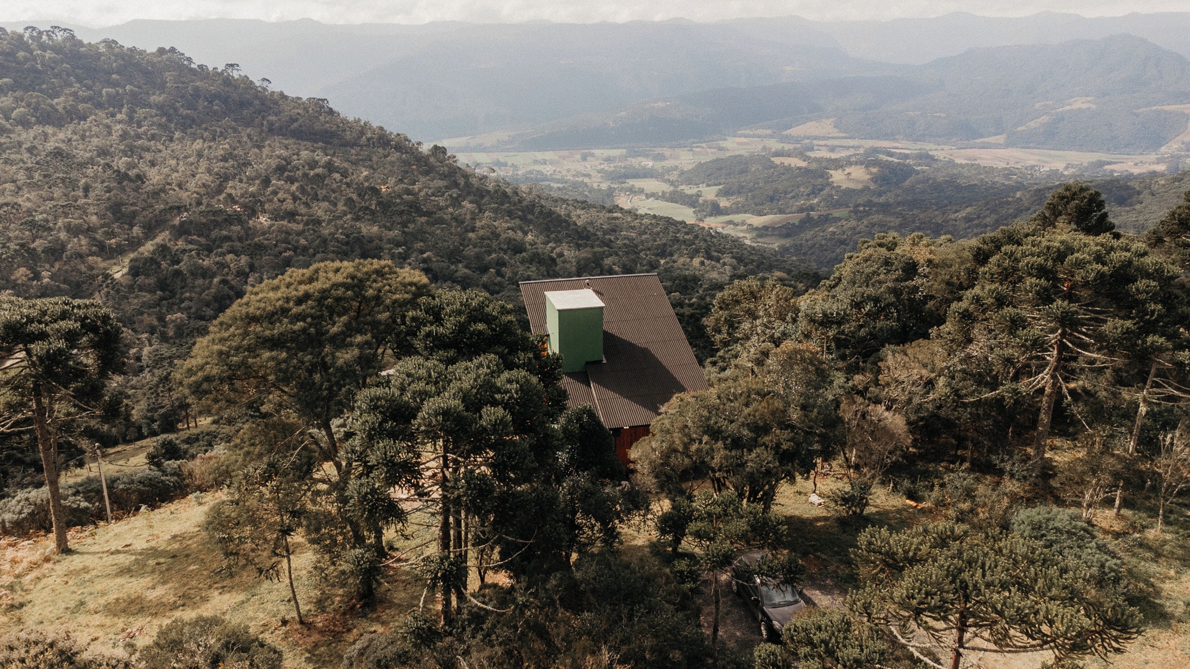 A remote cabin with a green chimney, surrounded by dense forest and hills.
