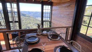 Un comedor con una mesa de madera dispuesta para cuatro, ventanas amplias que ofrecen vistas a las montañas y una lámpara colgante.