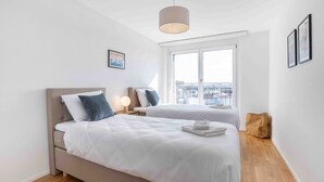 A modern hotel room with two beds, a large window with a city view, a hanging light fixture, and a framed picture on the wall. - Arsenaux Residence by Homenhancement (Fribourg)