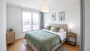 A modern bedroom with a large bed, a bedside table with a lamp, a window with curtains, and two wall art pieces. - Arsenaux Residence by Homenhancement (Fribourg)