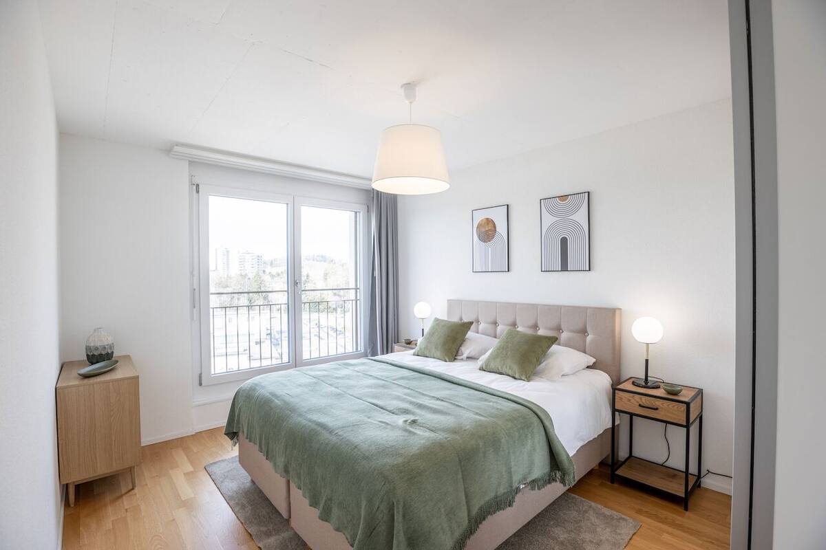 Un dormitorio moderno con una cama grande, una mesita de noche con lámpara, una ventana con cortinas y dos cuadros en la pared.