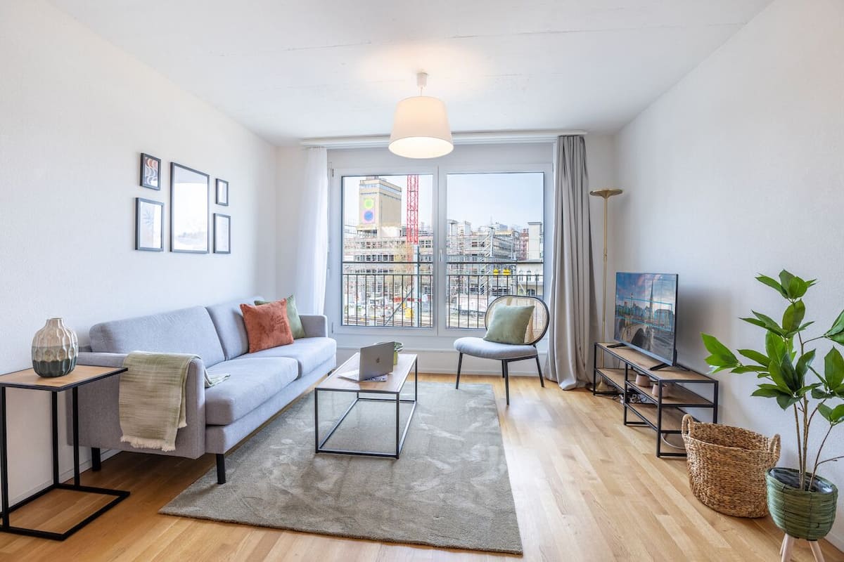 Una sala moderna con un sofá gris, una mesa de centro, un televisor sobre un mueble bajo y un ventanal con vistas a la ciudad.