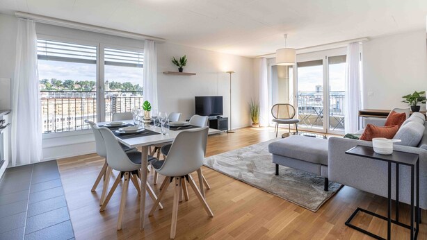 A modern living room with a dining area, a grey sofa, a TV, and a balcony with a view. - Arsenaux Residence by Homenhancement (Fribourg)