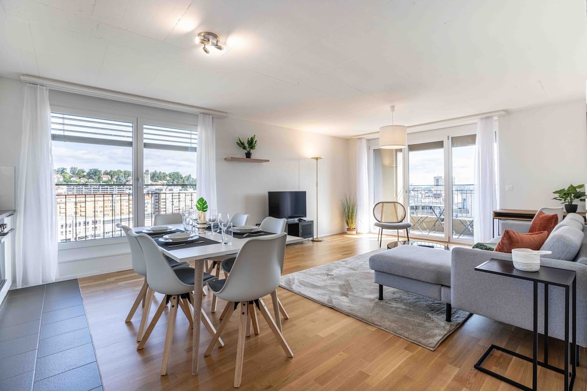 Una sala de estar moderna con comedor, un sofá gris, televisor y balcón con vistas.