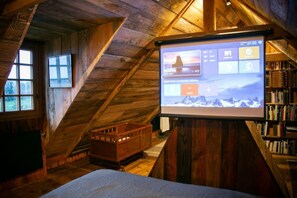 Una habitación cálida en el ático, con techo inclinado de madera, una cama, un cofre de madera y una pantalla de proyector mostrando un paisaje montañoso.