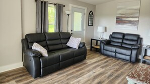 A living room with a black leather sofa, a black leather armchair, a wooden floor, a ceiling fan, and a door with a window. - Beautiful Country Setting Welcome to Moberly Manor (Depauw)