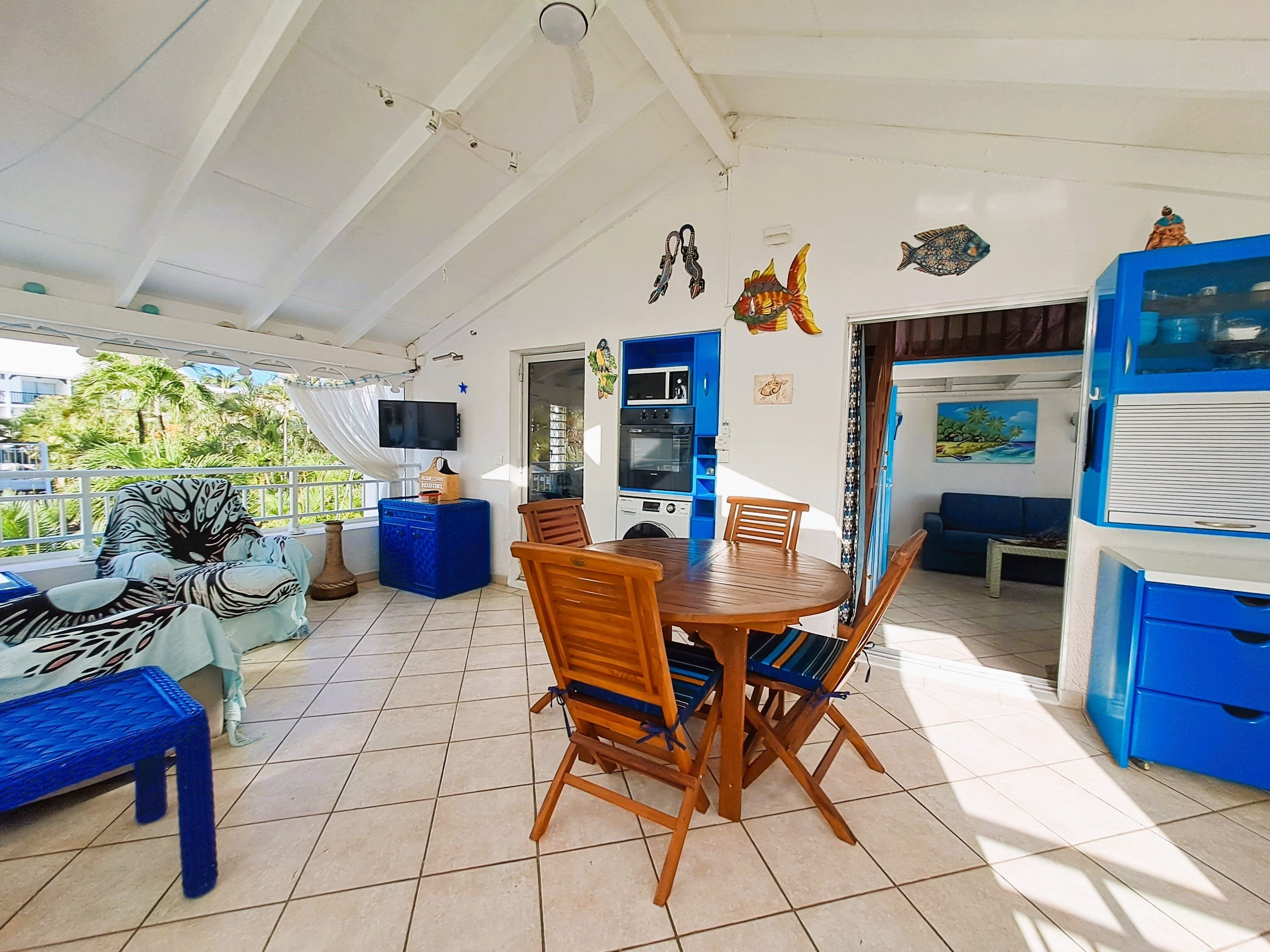 Ein überdachter Patio-Bereich mit Esstisch und Stühlen, einem blauen Sofa und einem an der Wand befestigten Fernseher.
