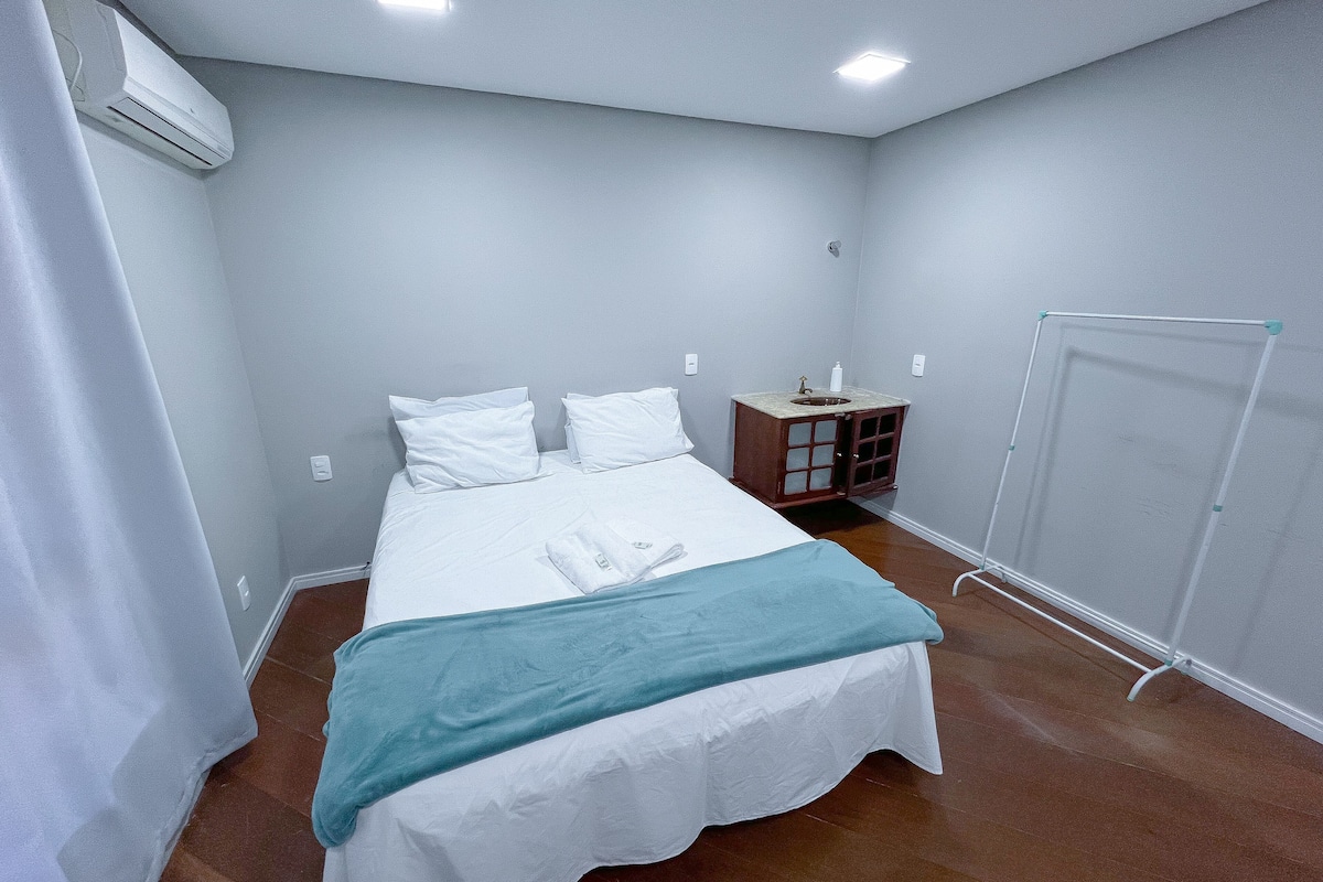 A neatly made bed with white bedding and a teal throw, a small wooden nightstand with a sink, and a standing mirror.