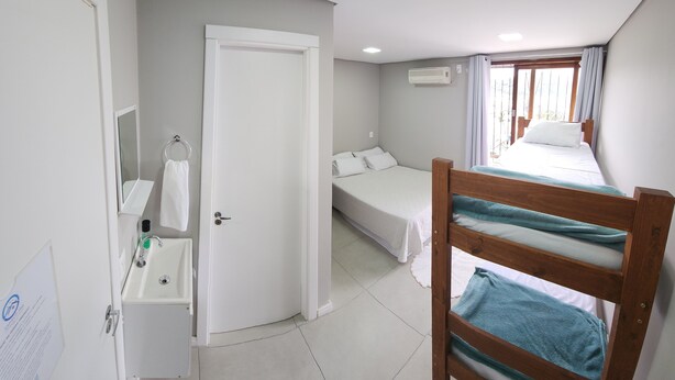 Quarto com uma cama de solteiro, uma cadeira de madeira com toalha, uma pia pequena e um espelho.