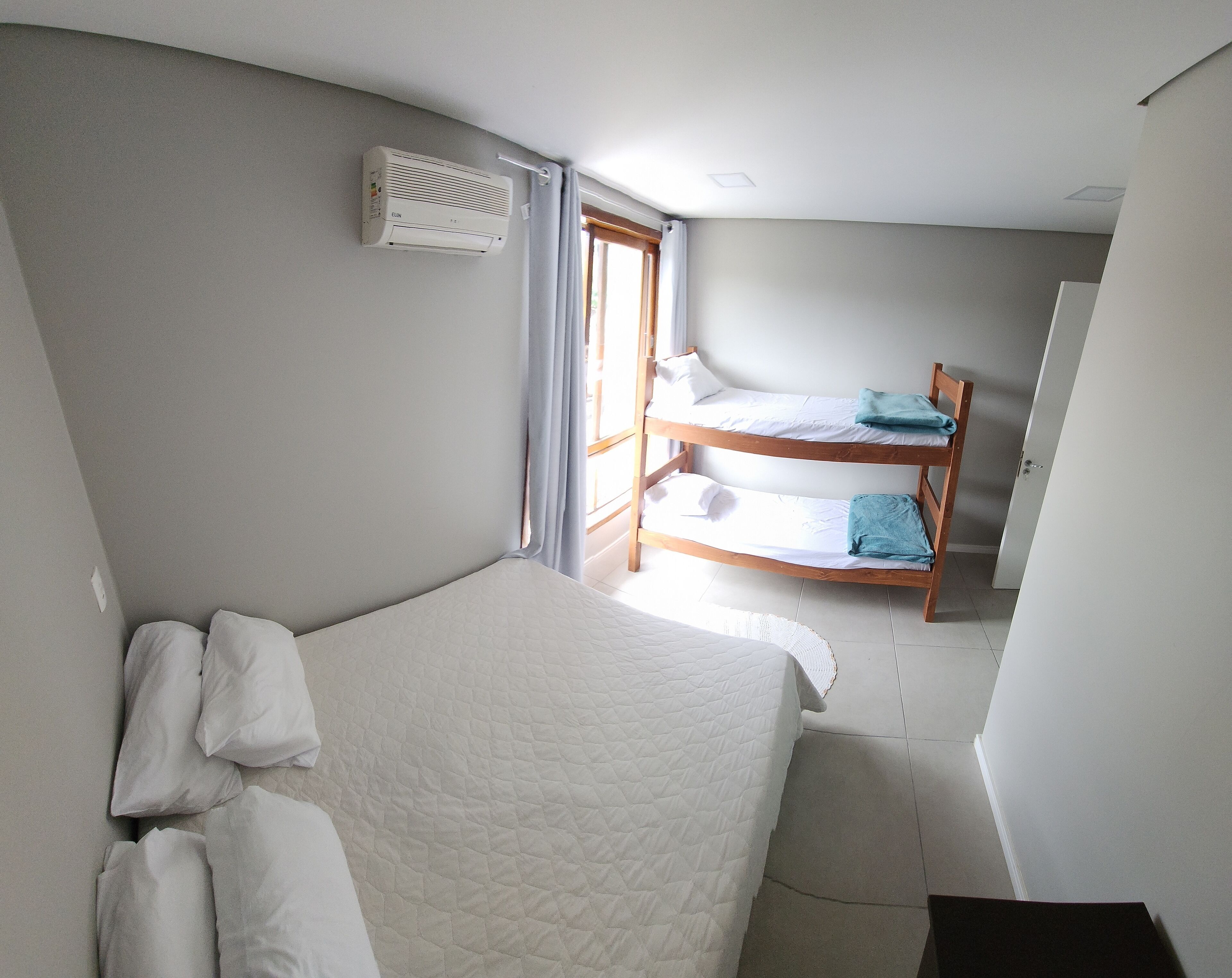 Una habitación con una cama individual, una cama litera al fondo, una ventana con cortinas y una rejilla en el techo.