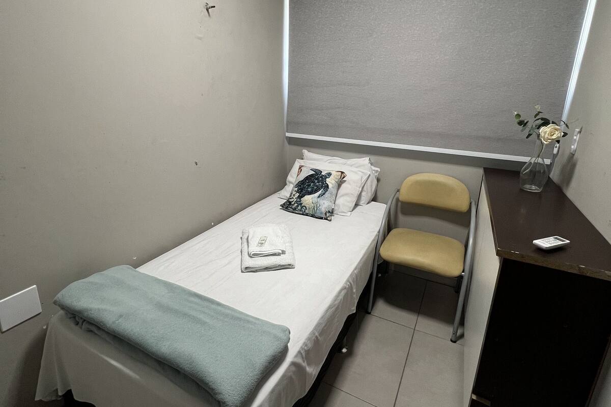 A small, neatly arranged room with a single bed, a desk, and a chair. An air conditioning unit is mounted on the wall above the bed.