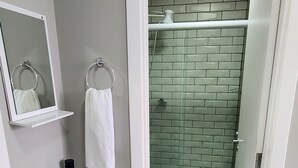 Casa de banho com lavatório branco, espelho e suporte para toalhas. Tem sanita e chuveiro com parede em azulejo verde.