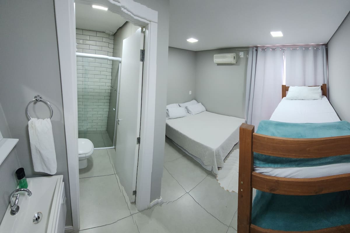 A small, clean room with a wooden bunk bed, a single bed, a bathroom with a toilet and shower, and a sink.