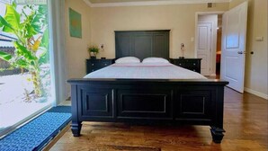 A bedroom with a large bed, a nightstand, a dresser, and a window with a view of greenery. - Carlsbad Beach Oasis+HotTub (Carlsbad)
