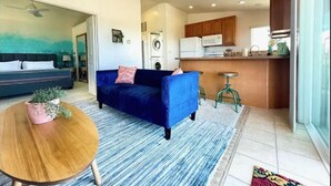 A modern living room with a blue sofa, a round wooden coffee table, and a kitchen area with wooden cabinets and a microwave. - Ocean View Home w/PrivateBalcony (Carlsbad)