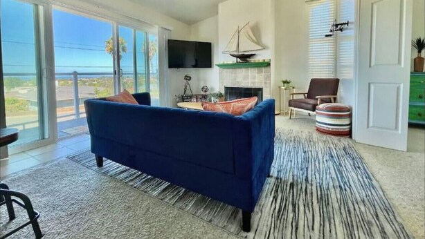 A modern living room with a blue sofa, a flat-screen TV, a fireplace, and a large window offering a view of the outdoors. - Ocean View Home w/PrivateBalcony (Carlsbad)