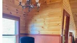 Een knus interieur met een houten plafond, een zwarte leren fauteuil, een tafeltje met een vaas en een raam met jaloezieën.