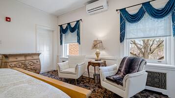 Une chambre d’hôtel avec un lit, deux fauteuils blancs, une petite table avec une lampe et une fenêtre avec des rideaux bleus.