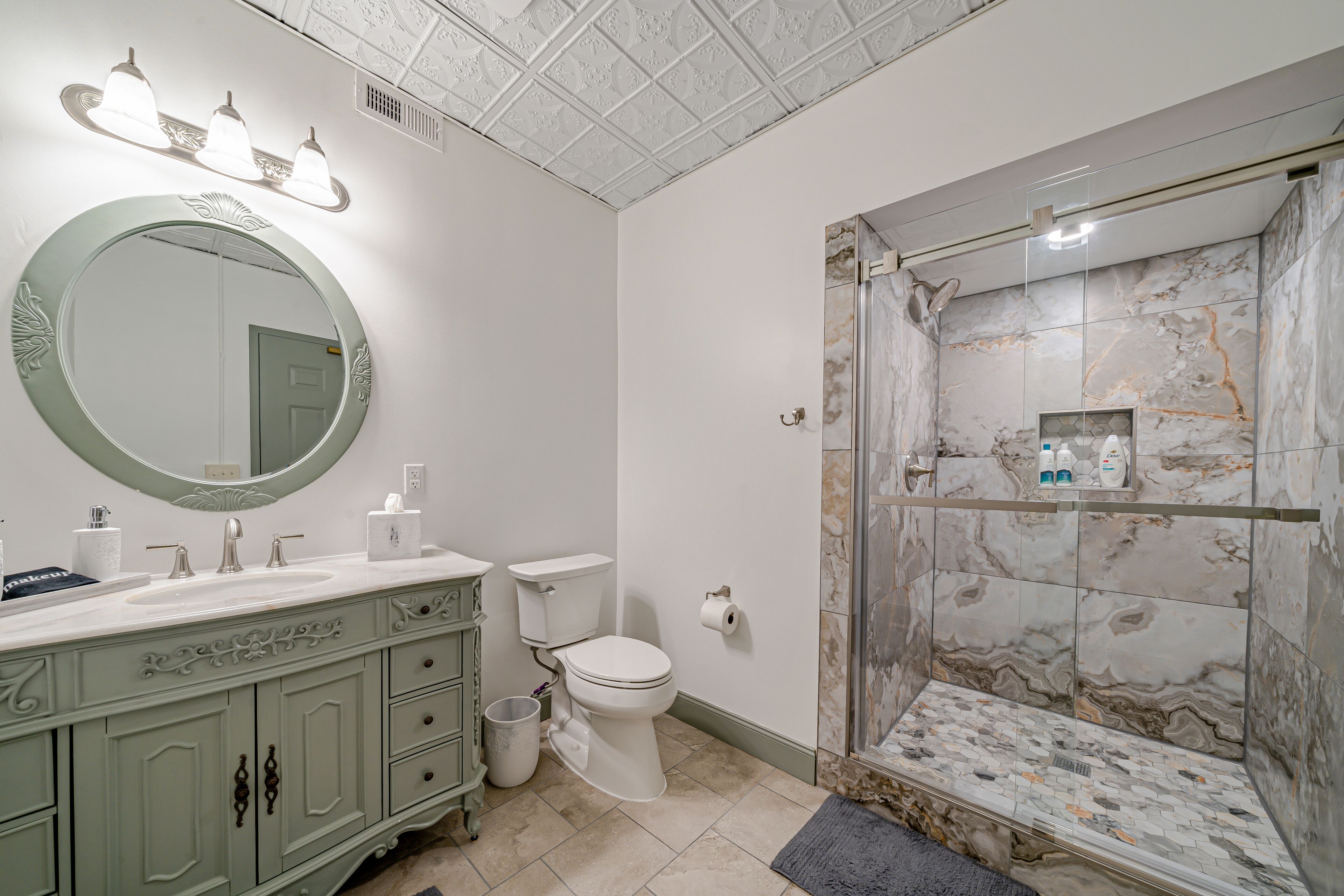 A bathroom with a marble shower, a round mirror, and a vanity with a sink and cabinet.