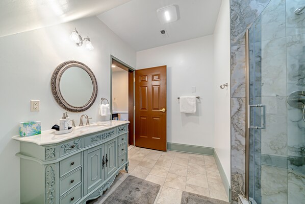 A bathroom with a marble shower, a vanity with a mirror, and a wooden door.