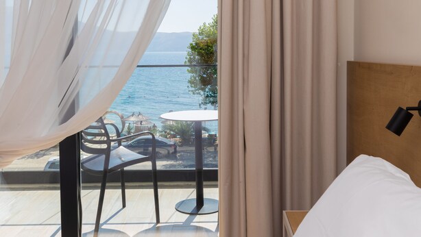 Une chambre à coucher avec un lit, une table de chevet et une vue sur la mer à travers de fines rideaux.