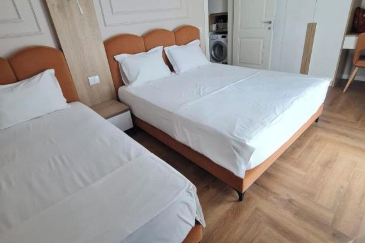 A bedroom with two beds, wooden flooring, a washing machine in the corner, and a door leading to another room.