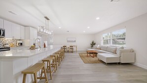 Una cucina moderna con isola centrale, sgabelli di legno, zona pranzo con tavolo e sedie, e area living con divano e ampie finestre.
