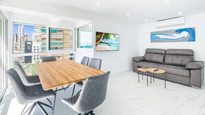 A modern living room with a wooden dining table, grey upholstered chairs, a grey sofa, and a small coffee table. There is a large window with a city view, a mounted TV, and an air conditioning unit on the wall.