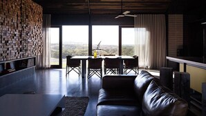 A modern living room with a brick wall, a large window, a dining table with chairs, and a leather sofa. - HILLPLAINS HOUSE - 100% OFF GRID LUXURY (Metcalfe)