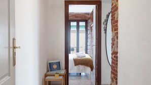 Een gang die leidt naar een slaapkamer met een bed, een kleine tafel met een plant en een ingelijst kunstwerk aan de muur.