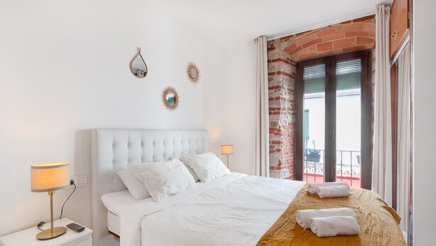 Een hotelkamer met een wit bed, een klein nachtkastje met een lamp, en uitzicht op een bakstenen gebouw door een raam.